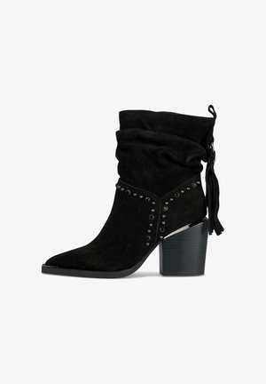 Zwarte suède enkellaars met een slungelige schacht, spitse neus, versierd met zilverkleurige studs, en een chunky hoge hak met kwastdetails.