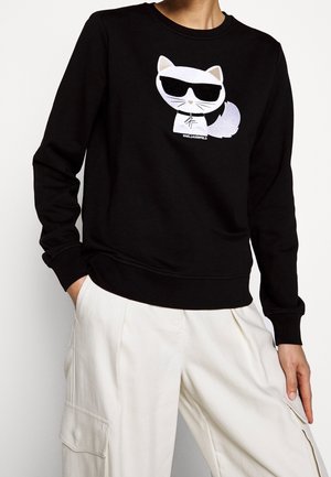 Person iført sort sweatshirt med hvid tegneserie kat med solbriller broderet på brystet, kombineret med løse hvide cargobukser.