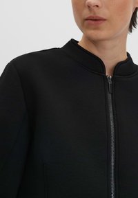 Veste zippée noire avec un col montant, fabriquée à partir d'un tissu lisse et texturé. Présente une fermeture éclair argentée et des accents de design minimalistes.