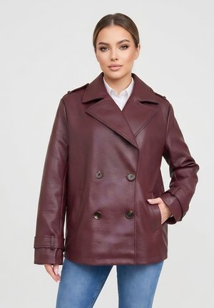 Kobieta z prostymi brązowymi włosami w bordowej, dwurzędowej skórzanej kurtce i niebieskich dżinsach, ręka w kieszeni, neutralne tło.