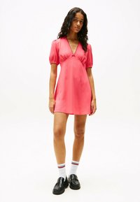 Vestido corto rosa con mangas abullonadas, escote en forma de V y detalles fruncidos. Combinado con zapatos negros y calcetines blancos con rayas rojas.