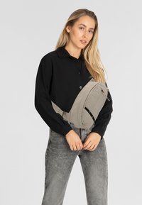 Borsa a tracolla grigio chiaro realizzata in un tessuto liscio con chiusura a zip e tracolla regolabile; indossata sopra una camicia nera con bottoni.