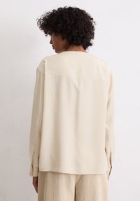 Helle beige, langärmlige Bluse mit lockerer Passform. Verfügt über eine gerade Rückennaht, geknöpfte Manschetten und eine weiche Stofftextur.