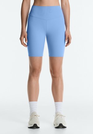 Frau trägt hellblaue, hoch taillierte Fahrradshorts, weiße Sportsocken und weiße Turnschuhe, steht vor weißem Hintergrund.