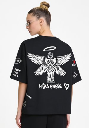Frau mit Dutt trägt ein schwarzes, übergroßes T-Shirt mit weißer Engel-Grafik und dem Text "Mira Paris" auf dem Rücken.