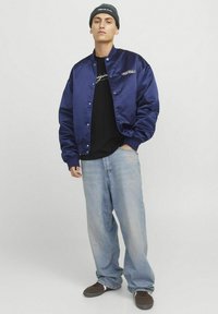 Mørkeblå satin bomberjakke med knaplukning, parret med en sort T-shirt og lyseblå baggy jeans. Sort hue med logo.
