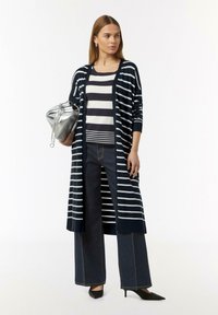 Langer navy Cardigan mit weißen Streifen über einem gestreiften Top, kombiniert mit weiten, dunklen Jeans und schwarzen, spitzen High Heels. Silberne Handtasche als Akzent.