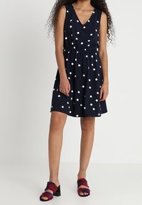 Granatowa, do kolan sukienka w białe kropki, zaprojektowana w serek, z elastycznym pasem i bez rękawów. Poł�ączona z kolorowymi, masywnymi sandałami.