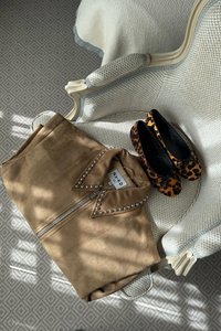 Kreemikattega faux suede jakk, millel on nupukesed krae, on eksponeeritud toolil, kõrval mustad ja pruunid leopardimustriga plätud tekstureeritud pinnal.