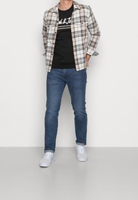 Camisa de cuadros en beige y gris sobre una camiseta negra, combinada con jeans azules y zapatillas blancas. Cuenta con cierre de botones en el frente y mangas con puños.