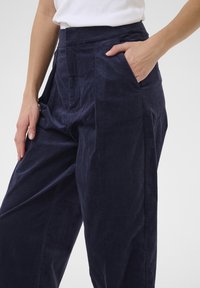 Pantaloni larghi blu navy realizzati in un tessuto morbido, dotati di tasche laterali, vita alta e dettagli plissettati sul davanti.