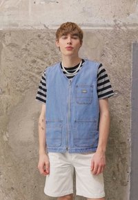 Denim ærmeløs vest i lys blå farve med to frontlommer og lynlås. Båret over en sort og hvid stribet skjorte og hvide shorts.