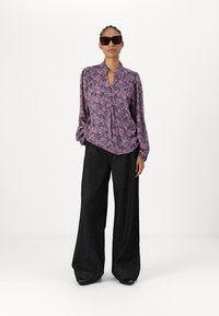Blusa viola a fantasia con colletto, maniche lunghe e vestibilità ampia abbinata a pantaloni neri a gamba larga. Occhiali da sole neri completano il look.