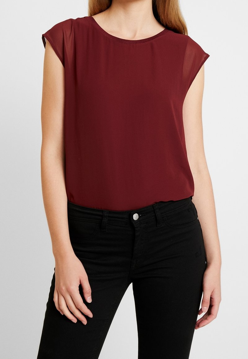 Femme portant un chemisier sans manches rouge foncé rentré dans un pantalon noir taille haute avec un bouton et des passants pour ceinture.