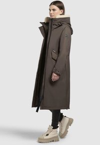 Lang brun frakke med høj krave og lammeskindsfor, som har en lynlås foran, sidelommer og et stilrent, minimalistisk design.