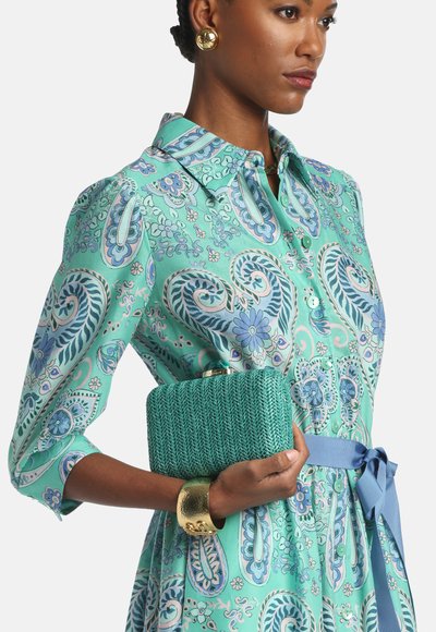 Turkis vævet clutch med rektangulært oblik og guldlås; båret med en mønstret grøn kjole og guldbelagt manchetarmbånd.