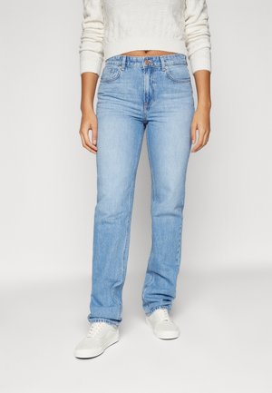 Jeans de mezclilla azul claro con un ajuste recto, lazos para el cinturón, bolsillos frontales y un cierre de un solo botón. Combinados con zapatillas blancas.