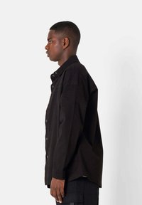 Black button-up shirt with a relaxed fit, featuring long sleeves, a pointed collar, and a pocket detail on the chest. Smooth fabric texture.