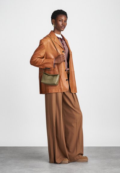 Bruine leren blazer met een relaxte pasvorm, gecombineerd met brede beige broeken. Een kleine olijfgroene tas hangt van de schouder. Subtiele textuurdetails.