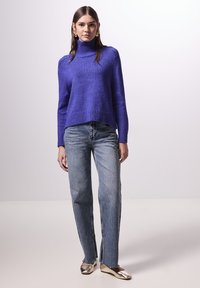 Paarse gebreide coltrui met ribbelzoom, gecombineerd met lichtblauwe high-waisted recht gesneden jeans en metallic gouden loafers.