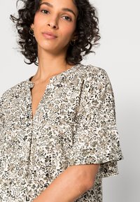 Blusa de estampado floral em tecido leve, com decote em V, mangas curtas com volantes e um fundo bege claro com detalhes em verde e castanho.