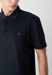 Homme portant un polo bleu marine foncé à manches courtes avec trois boutons et un petit logo circulaire sur la poitrine gauche.