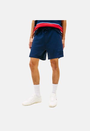 Mann trägt marineblaue Shorts mit Kordelzug, weiße Socken, weiße Sneakers und einen gestreiften Pullover in Rot, Blau und Schwarz.