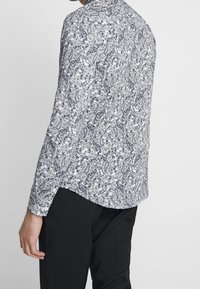 Chemise à manches longues en motif paisley noir et blanc, fabriquée en coton. Dispose d'un ourlet arrondi et de poignets à boutons. Design ajusté.