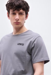 Νέος άνδρας με καστανά σγουρά μαλλιά, φοράει γκρι μπλουζάκι Levi's, κοιτάζει κατευθείαν μπροστά με φόντο μια απλή λευκή επιφάνεια.