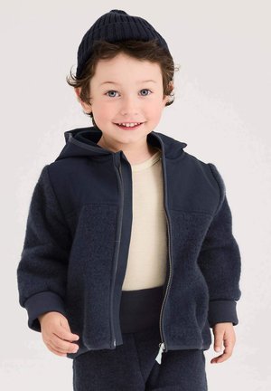 Marineblaues Zip-up-Jacke mit Fleece- und glatten Einsätzen, ausgestattet mit einer Kapuze und gerippten Bündchen. Getragen über einem hellbeigen Hemd und passenden Hosen.