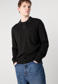 Schwarzes, gestricktes Poloshirt mit Kragen, zwei Knöpfen und gerippten Bündchen. Verfügt über dezente Markierung und einen lässigen Schnitt, kombiniert mit blauen Jeans.