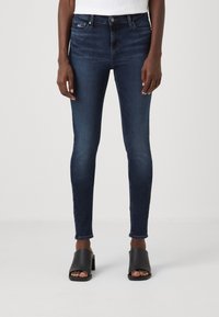 Jeans ajustados de mezclilla en lavado oscuro, con una cintura alta, bolsillos frontales y desvanecimientos sutiles. Combinados con sandalias de tira abiertas negras.