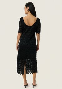 Mustaklainen pitsimekko l短ineillä hihoilla, syvällä avomahaalilla, aaltoilevalla helmanreunalla ja sivureiällä. Sujuva rakenne ja monimutkainen kukkakuosi.