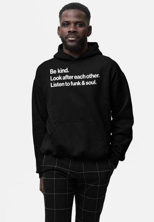 Homme avec barbe portant un sweat à capuche noir avec le texte : « Soyez gentils. Prenez soin les uns des autres. Écoutez du funk et de la soul. » et un pantalon noir à lignes blanches en damier.