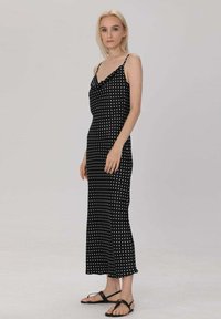 Robe noire à pois avec un col drapé, des bretelles spaghetti et une silhouette ajustée, faite en tissu léger ; modèle portant des sandales plates noires.