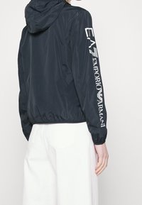 Marinblå huva vindjacka med elastiska ärmslut och nederkant. Har vit logotyp längs ärmen. Slät, lätt material.