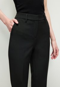 Zwarte op maat gemaakte broek van een soepele stof, met twee zijzakken en een vlakke voorkant. Slim-fit ontwerp met subtiele plooien langs de benen.