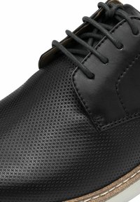 Scarpa in pelle nera con tomaia perforata, design con lacci, suola di un marrone chiaro a contrasto e accenti cuciti. Finitura testurizzata visibile.