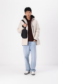 Beige gesteppte Jacke mit Kapuze, schwarzem Reißverschluss und zwei Taschen. Brauner Pullover und hellblaue Jeans. Schwarze Umhängetasche. Weiße Sneakers.