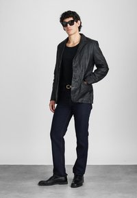 Schwarze Lederblazer mit einer zerknitterten Textur, getragen über einem schwarzen Hemd. Kombiniert mit dunklen Jeans und schwarzen Stiefeln, mit einem metallischen Gürtel.