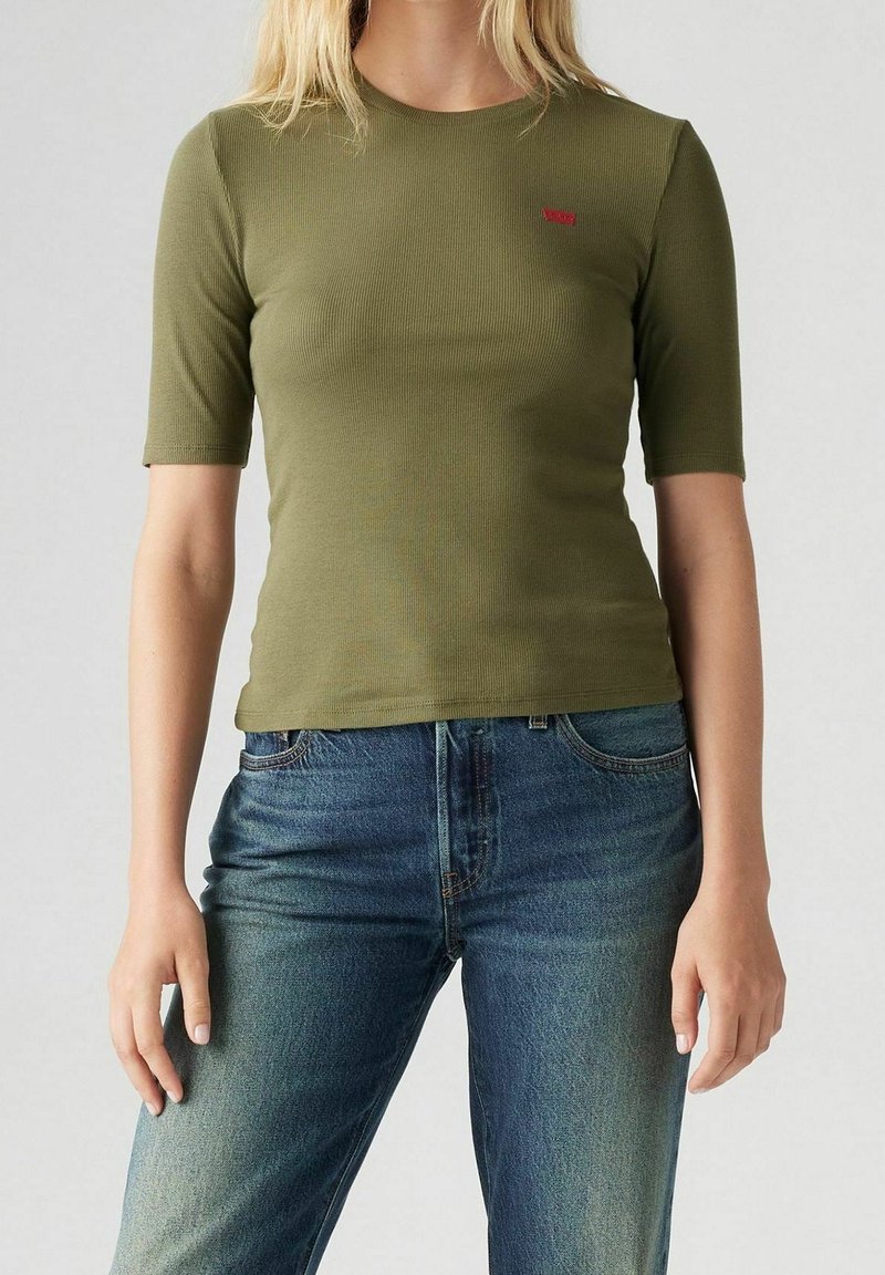 Femme portant un haut court à manches courtes côtelé vert olive et un jean en denim bleu, se tenant devant un fond uni.