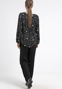 Blouse noire à motif d'étoiles blanches, manches longues et texture froncée. Associée à un pantalon noir ample et des chaussures noires.