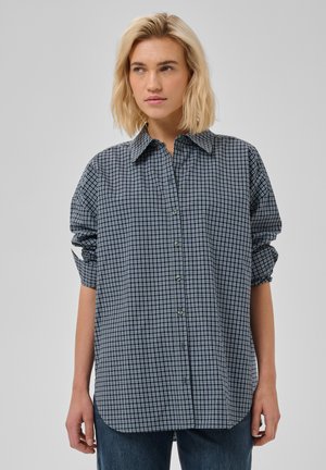 Vrouw met schouderlang blond haar draagt een loszittend blauw-wit geruit overhemd met knopen en een donkere spijkerbroek, staand tegen een effen achtergrond.