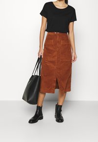 Žena v černém tričku, hnědé manšestrové midi sukni s předním rozparkem, černých kotníkových botách a nese velkou černou kabelku.