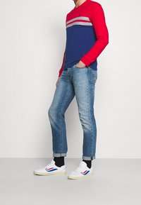 Maglietta a maniche lunghe rossa e blu con strisce grigie, abbinata a jeans blu chiaro e sneakers bianche con accenti rossi e blu.