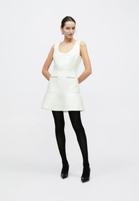 Biała sukienka bez rękawów z fakturowanej tkaniny, z rozkloszowanym dołem i dwoma przednimi kieszeniami. Zestawiona z czarnymi rajstopami i szpilkami.