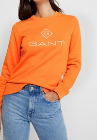Felpa arancione con colletto tondo e logo bianco "GANT". Abbinata a jeans a vita alta di colore blu chiaro, che mostrano uno stile casual.