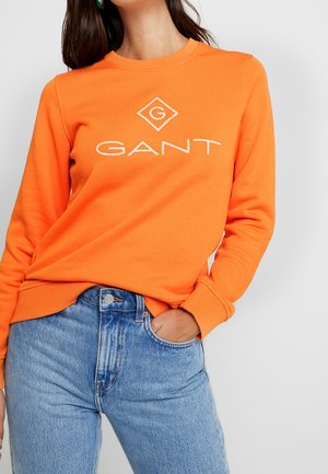 Sweat-shirt orange à col rond avec un logo "GANT" blanc. Associé à un jean taille haute bleu clair, représentant un style décontracté.