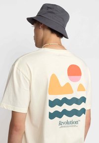 Camiseta de manga corta beige con un estampado gráfico en la parte posterior: montañas, sol y líneas onduladas en naranja, rosa y verde oscuro.