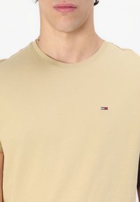 Beige pamut póló kerek nyakkivágással és rövid ujjal, melynek mellkasán egy kis hímzett logó található piros, fehér és kék színekben.